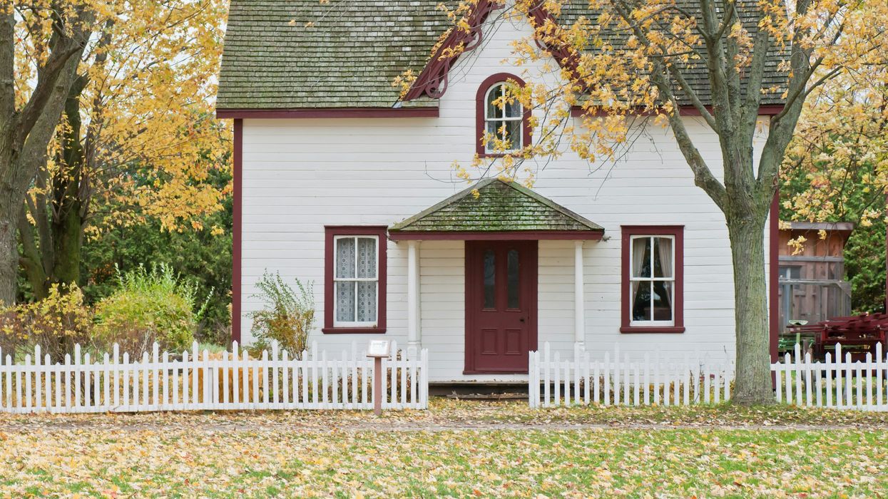 Charming white house with red trim, surrounded by autumn trees and a white picket fence.