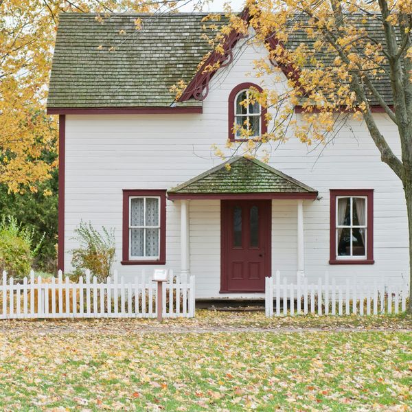 Charming white house with red trim, surrounded by autumn trees and a white picket fence.