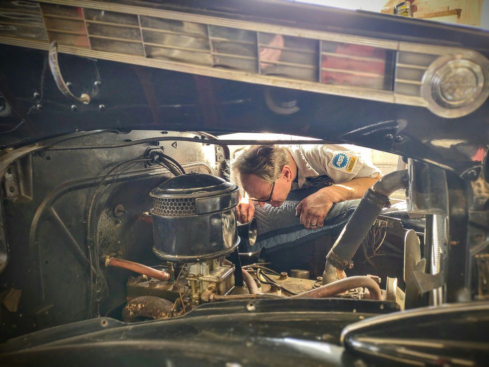 Mechanic inspecting an engine bay through the open hood of a vintage car.