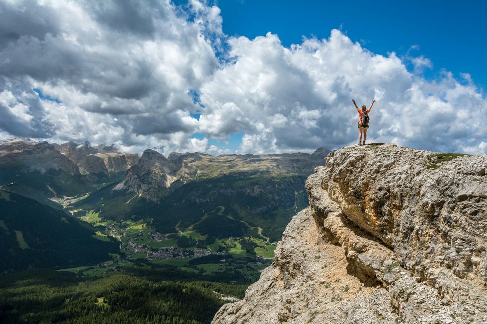 Person raising arms atop a rocky cliff with mountains and clouds in the background.