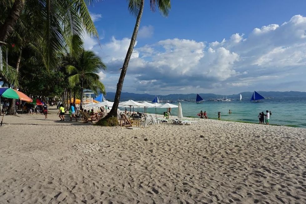 Sandy beach with palm trees, chairs, and sailboats on the blue sea under a cloudy sky.