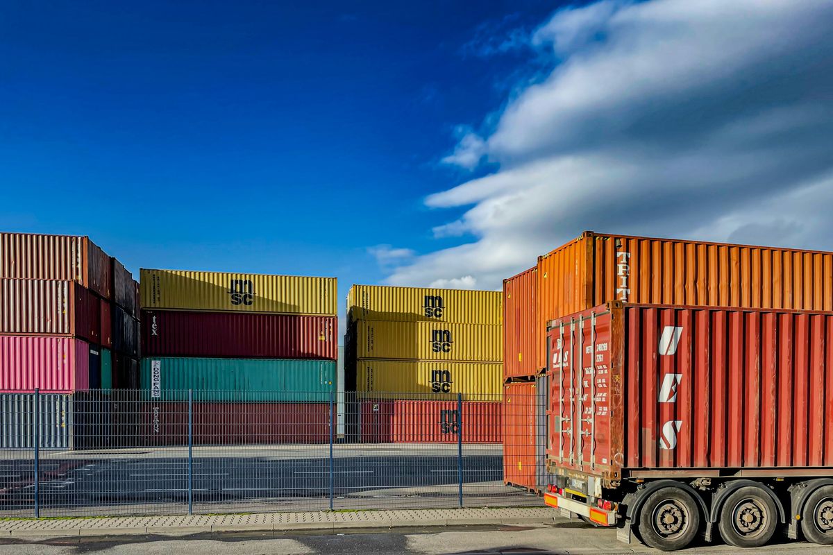 Stacked shipping containers and truck trailer under a blue sky with clouds.