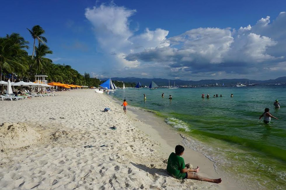 Sunny beach with palm trees, sailboats, and people swimming in clear blue water.