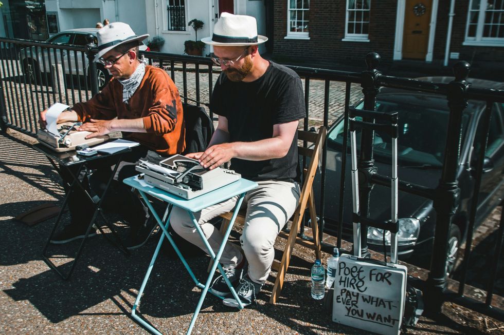 two poets typing on typewriters outdoors, and a sign reads "poet for hire, pay what you want"